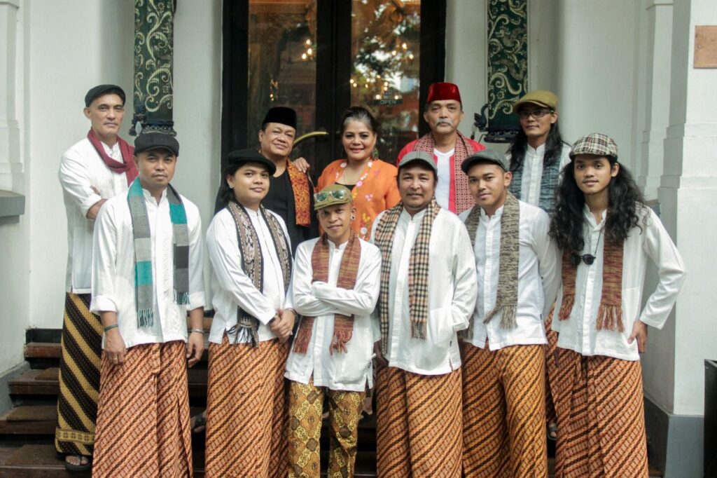 Whatsapp image 20260612 at 15 16 20 catrawarta Group of ten people posing on steps in traditional indonesian attire white shirts with batik sarongs and scarves some wearing hats or headgear outdoors in front of a doorway